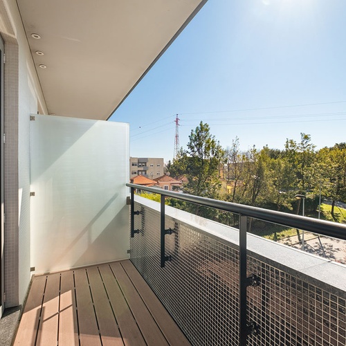 Uma varanda moderna e ensolarada, com piso de madeira, painel de vidro fosco e guarda-corpo metálico, oferece uma vista para árvores, edifícios e uma torre de alta tensão sob um céu azul.