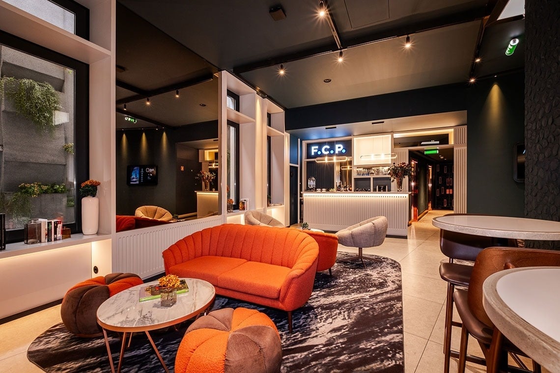 Um lounge moderno e acolhedor, possivelmente de um hotel, com sofás laranjas vibrantes, um bar elegante, teto escuro e um tapete estampado, criando um ambiente sofisticado.