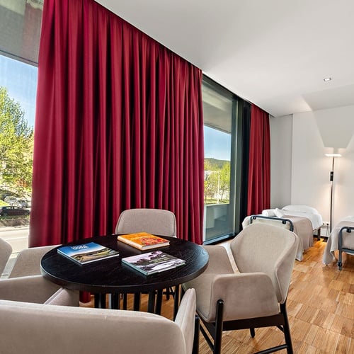 Um quarto luminoso apresenta uma grande janela com cortinas vermelhas, uma mesa redonda com cadeiras no centro e duas camas individuais encostadas à parede branca.