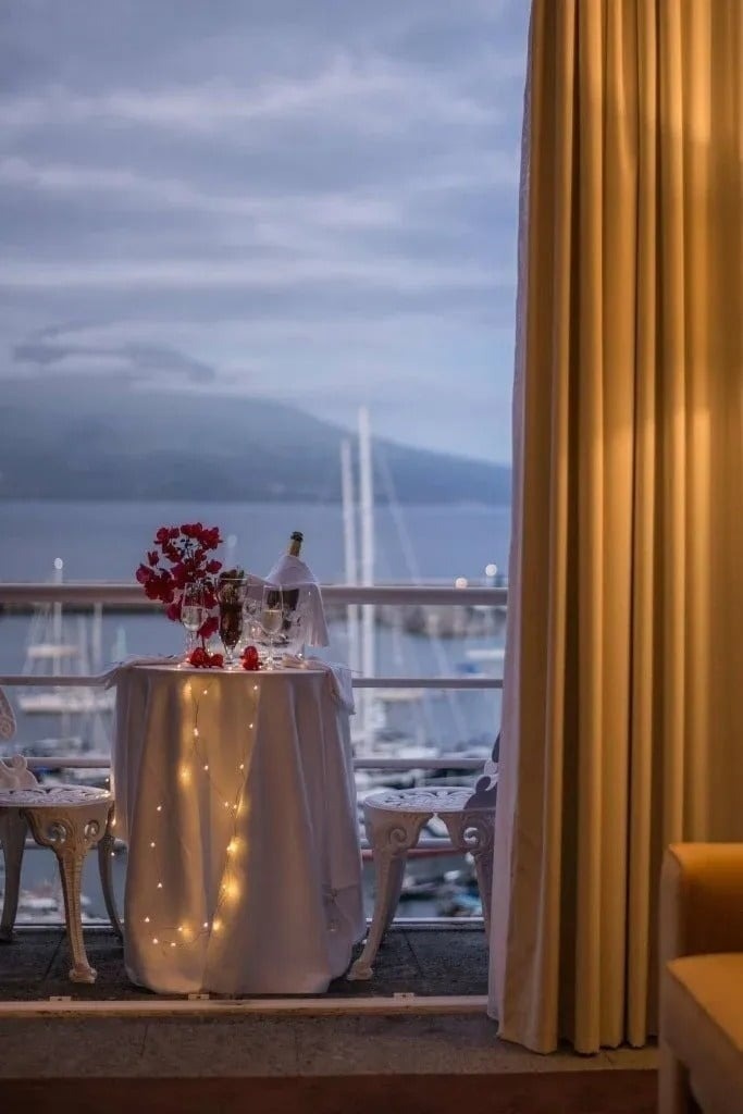 Uma mesa romanticamente posta com luzes de fadas, flores e champanhe, em uma varanda com vista para uma baía com barcos e montanhas sob um céu nublado.