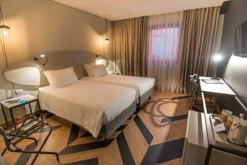 Um quarto aconchegante com roupa de cama branca e cortinas escuras oferece uma vista para o exterior através de uma grande janela, revelando árvores verdes e uma varanda.