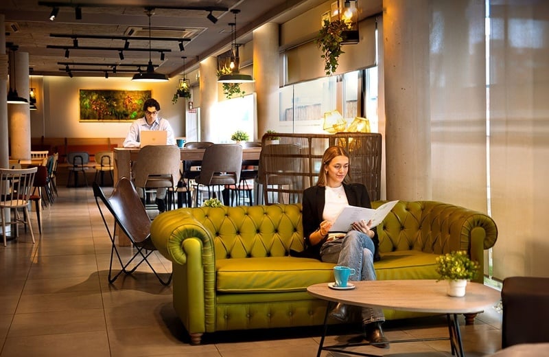 En un espacio de co-working moderno, un hombre trabaja en una laptop mientras una mujer lee en un sofá verde.