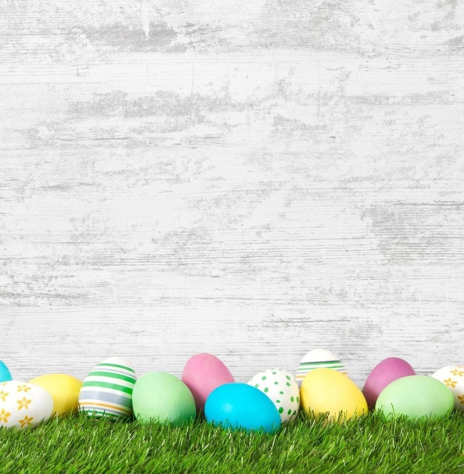Farbige Ostereier liegen auf grünem Gras vor einem weiß getünchten Holzhintergrund.