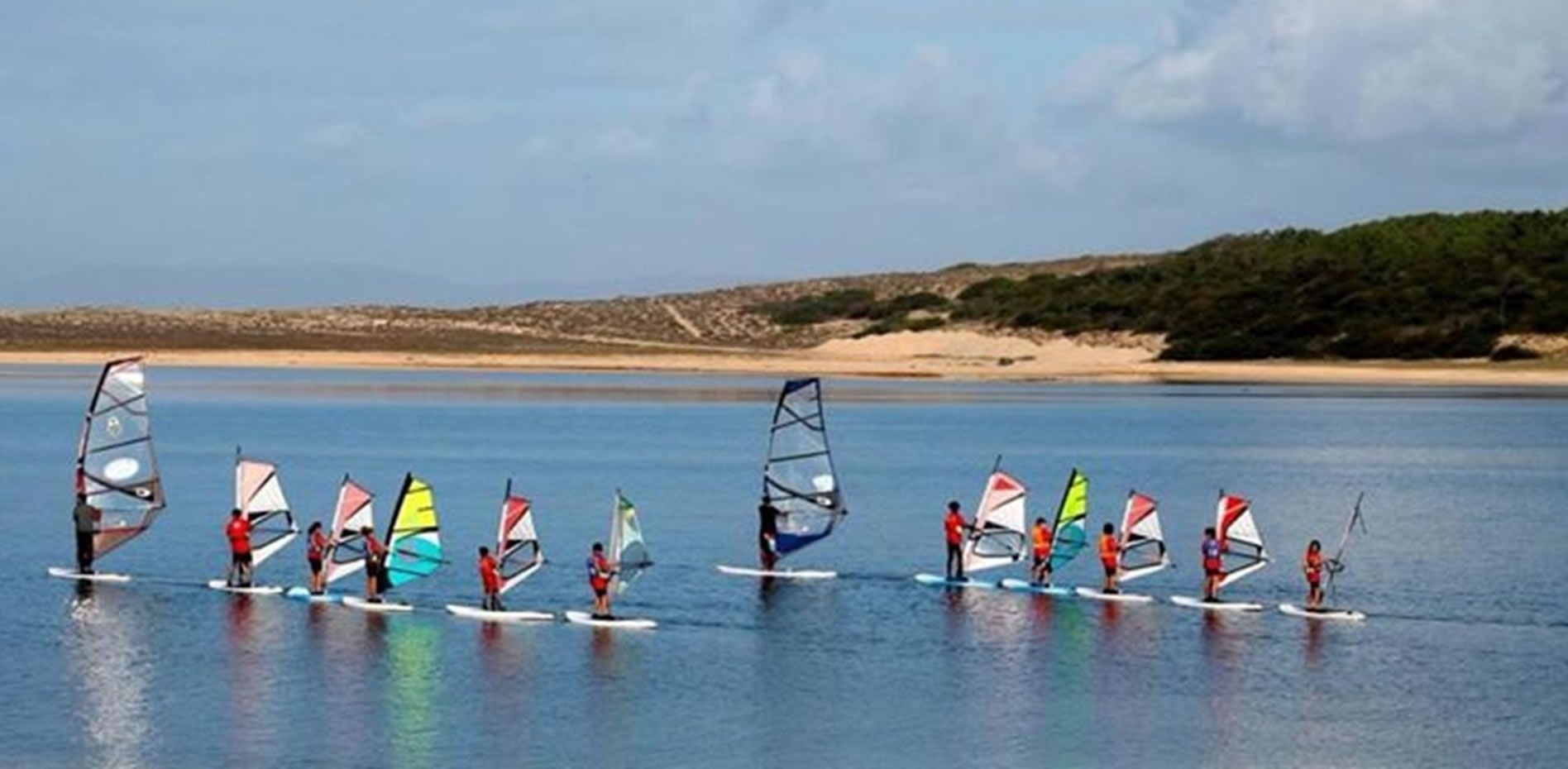 Um grupo de windsurfistas com velas coloridas navega em águas calmas, com dunas e vegetação costeira ao fundo.