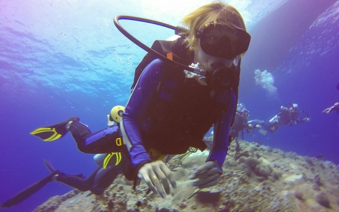 Uma pessoa mergulha com equipamento de scuba e máscara sob a superfície da água azul, com outros mergulhadores e um recife de coral ao fundo.