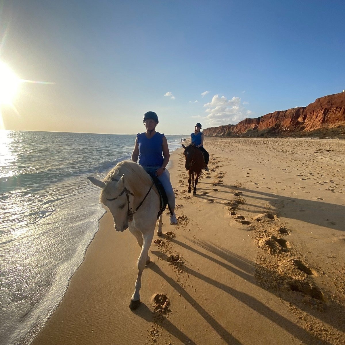 Duas pessoas cavalgam ao longo de uma praia ensolarada, ladeada pelo oceano à esquerda e por falésias rochosas e avermelhadas à direita.