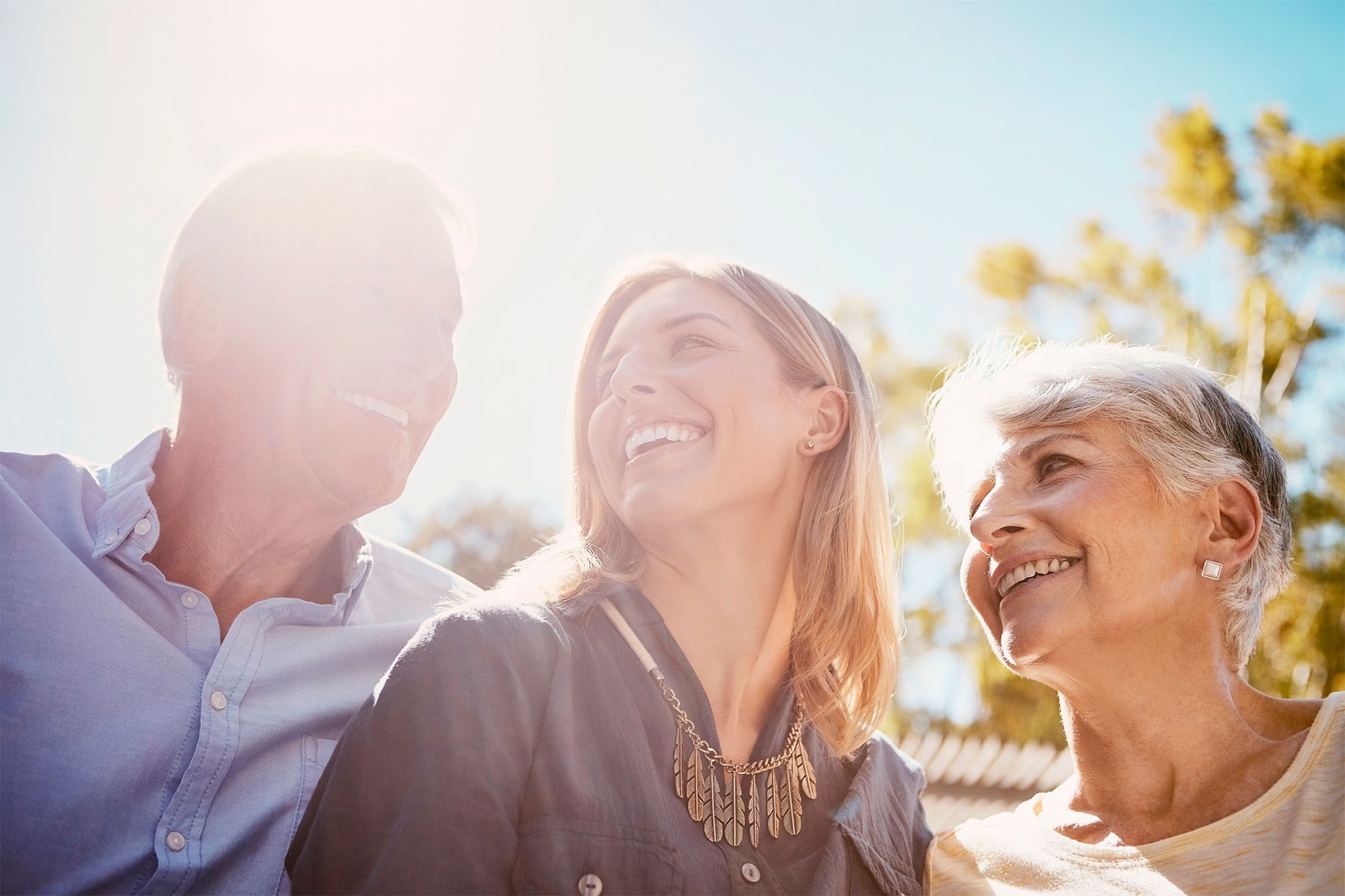 Tres personas sonrientes de diferentes generaciones miran hacia arriba o entre sí, bañadas por la luz del sol al aire libre.