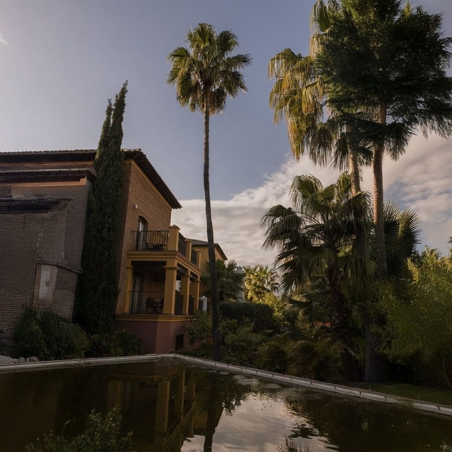 Un edificio de ladrillo con balcones, rodeado de palmeras y otros árboles, se alza junto a un estanque oscuro que refleja el cielo parcialmente nublado.