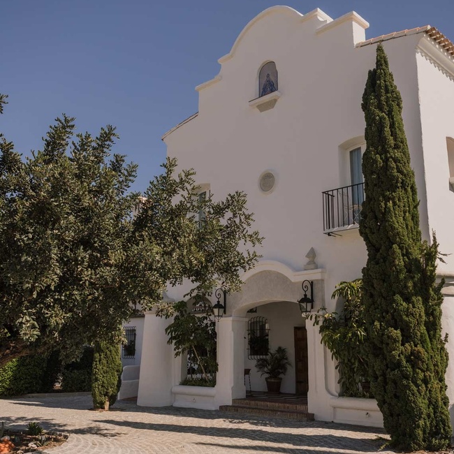 A serene outdoor swimming pool is set against a backdrop of a grand white building, tall cypress trees, and distant mountains under a bright, clear sky.
