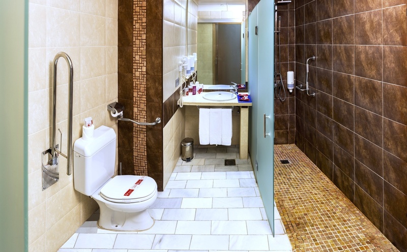 This modern and accessible bathroom features a toilet with grab bars, a sink, and a walk-in shower, all decorated with contrasting beige and brown wall tiles and light floor tiles.