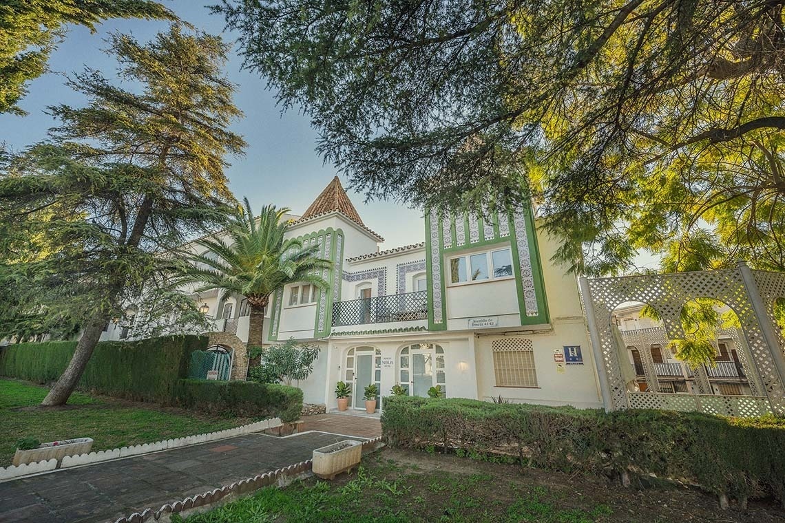 Un pintoresco hotel de fachada blanca con detalles decorativos en verde y azul, enmarcado por exuberante vegetación y altos árboles.