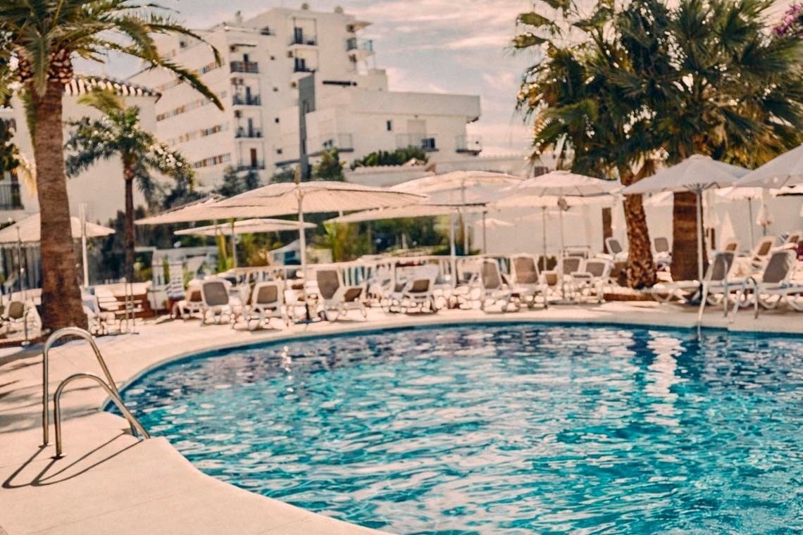 Una piscina exterior con tumbonas, sombrillas y palmeras, con un edificio blanco de fondo, bajo un cielo luminoso.
