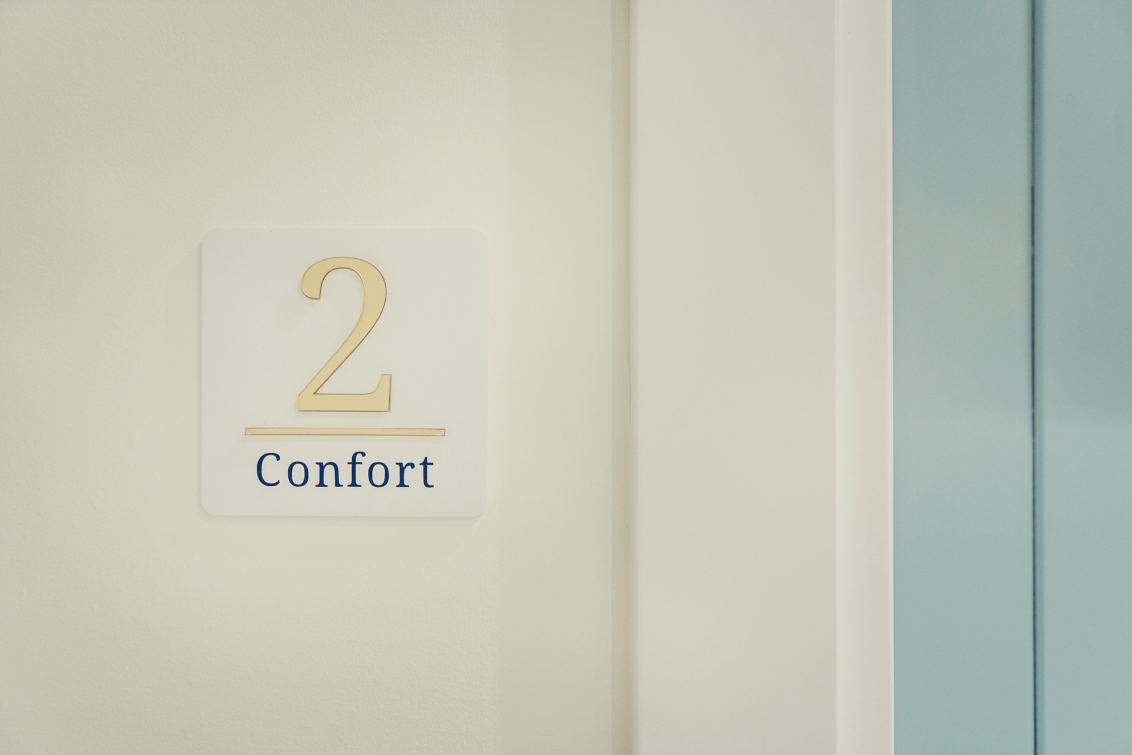 Un letrero blanco con el número dos en dorado y la palabra "Confort" en azul está en una pared clara, junto a un marco de puerta blanco y una superficie azul claro.