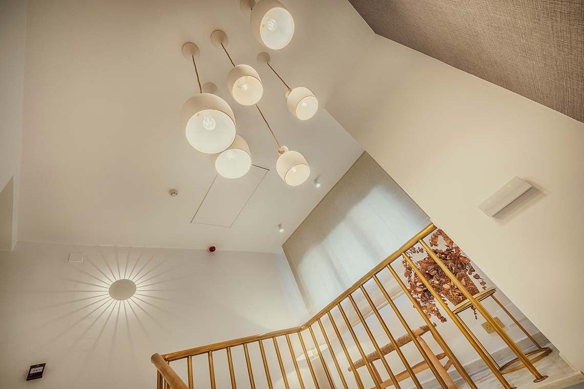 La imagen muestra el interior de una escalera con barandilla dorada, iluminada por un conjunto de modernas lámparas colgantes en el techo y una lámpara de pared decorativa con efecto de rayos de luz.