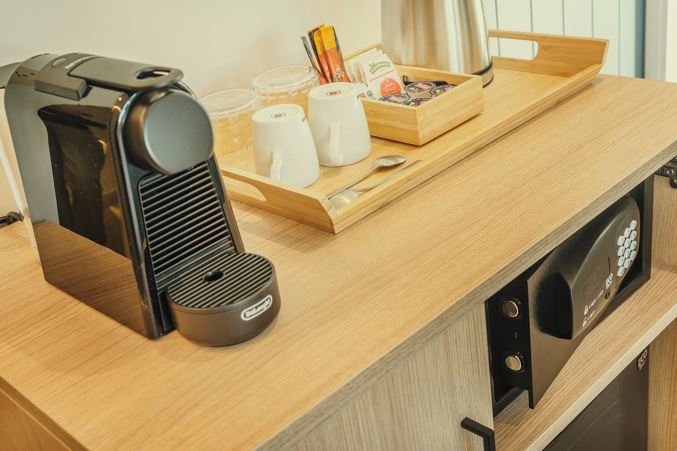 Una vista de un mostrador de madera con una máquina de café Nespresso, una bandeja de madera con tazas, un hervidor y artículos de té, y una caja fuerte empotrada debajo.