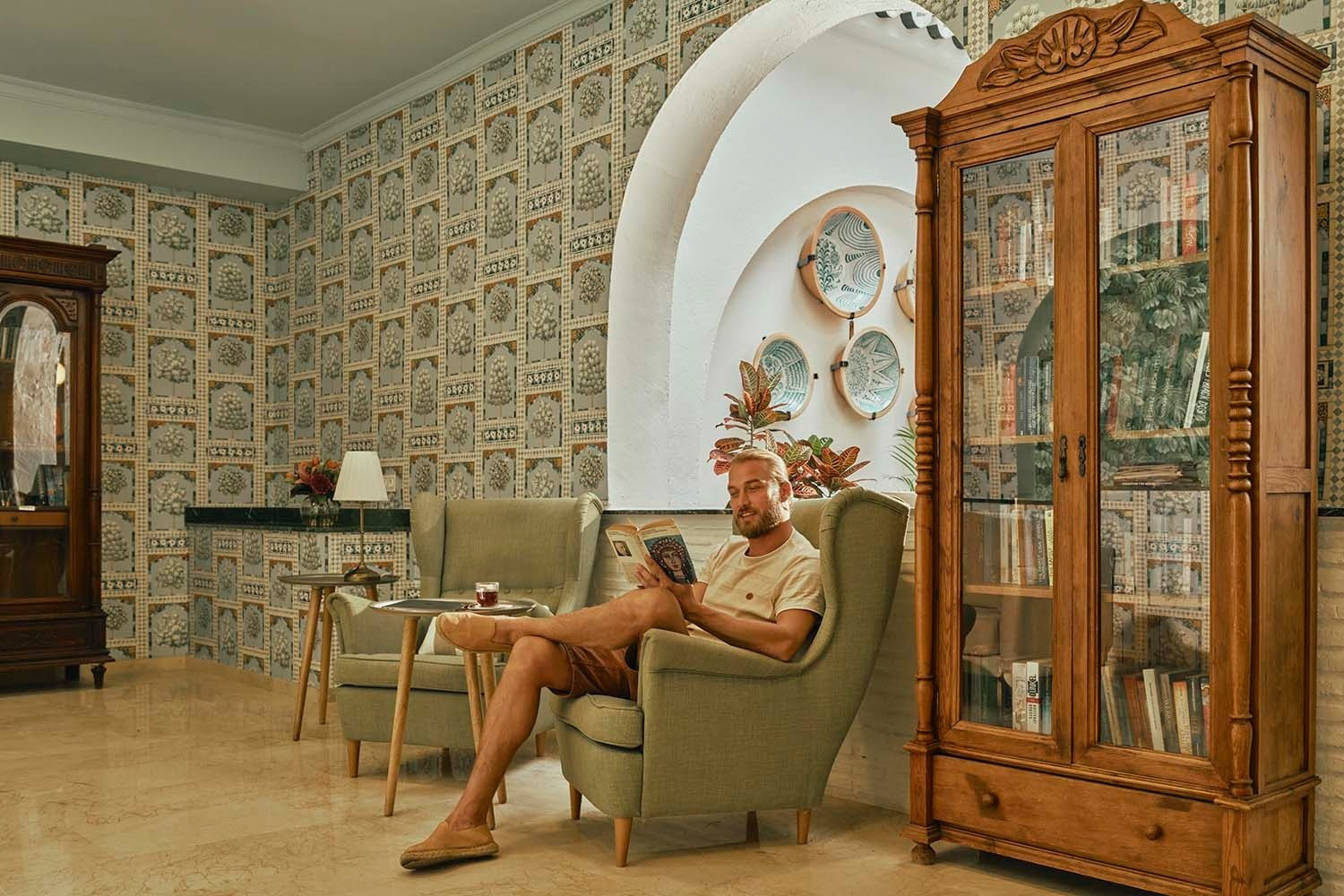 Villa Flamenca | Beneste Hoteles: Hombre leyendo en sillón verde junto a vitrina de madera.