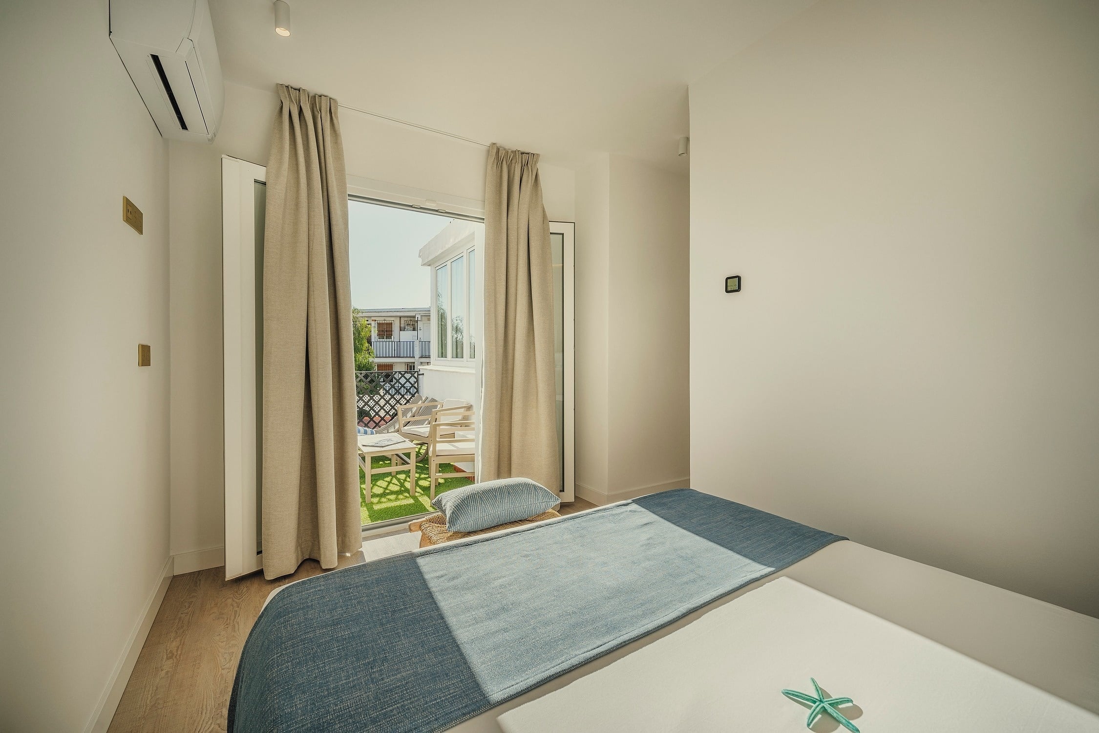 Un dormitorio moderno y luminoso con cama, decoración azul, suelo de madera y salida a un balcón con césped artificial y mobiliario exterior.