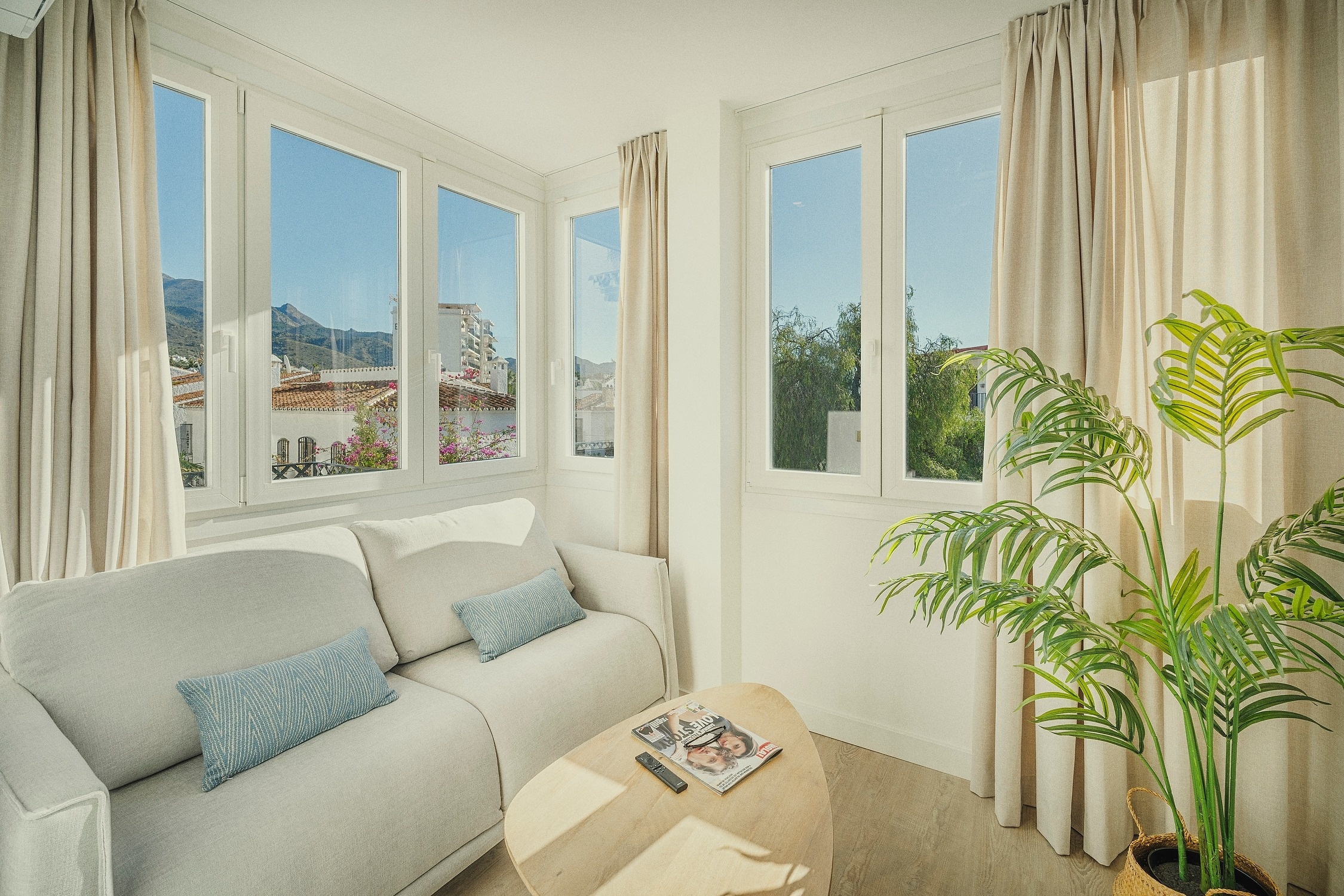 Un luminoso salón con un sofá blanco, una planta y grandes ventanales que ofrecen vistas a montañas y edificios.