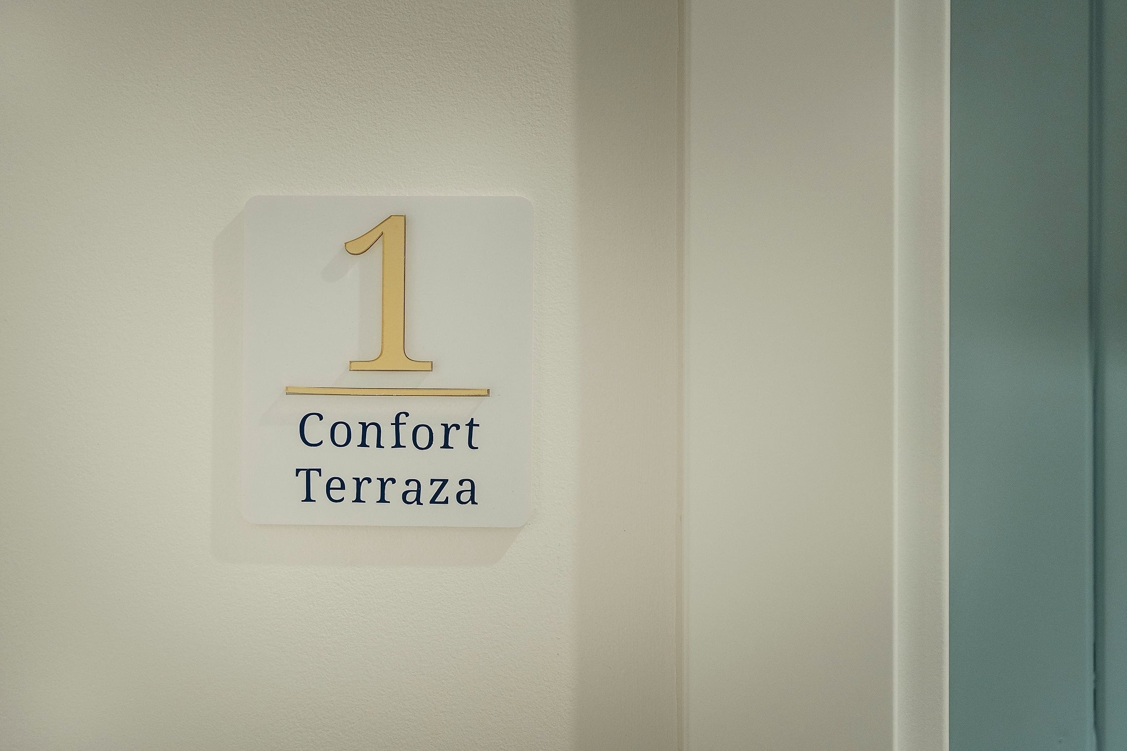 Un letrero blanco con un número 1 dorado y la inscripción "Confort Terraza" en azul está fijado a una pared de color claro, junto a un marco de puerta blanco y una puerta azul verdoso.