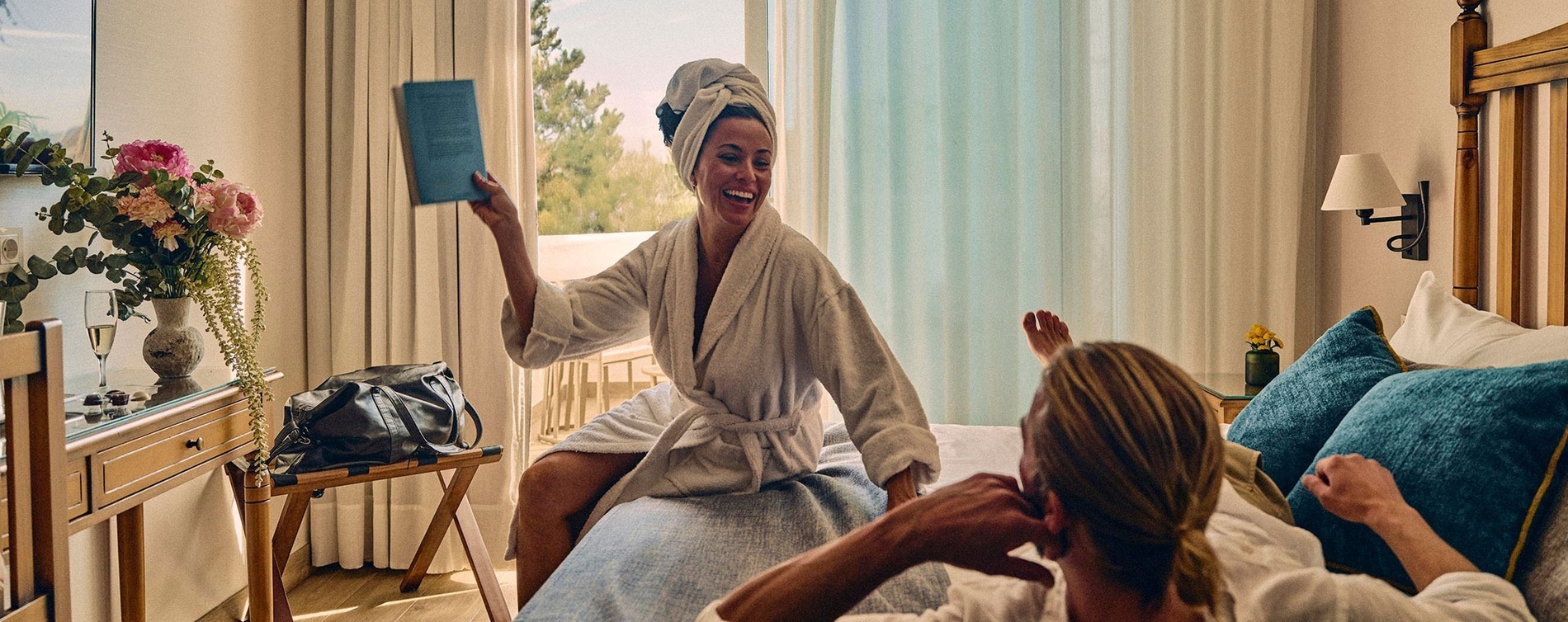 Una mujer en bata de baño con una toalla en la cabeza se ríe mientras sostiene un libro azul, compartiendo un momento alegre con otra persona en una habitación de hotel con flores y champán.