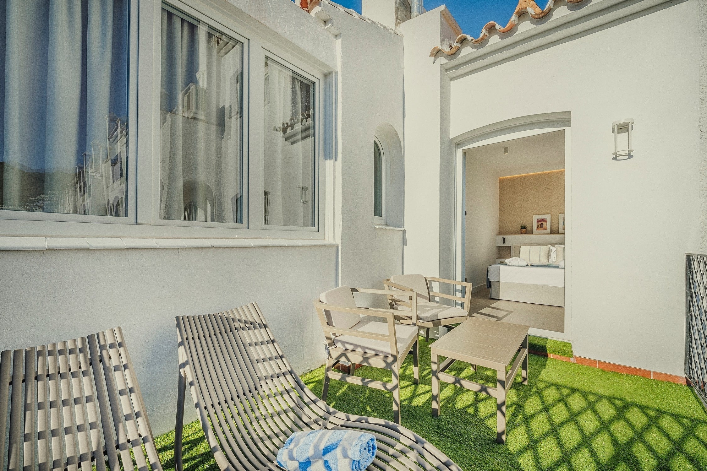 Una soleada terraza con césped artificial y mobiliario de exterior conecta con un dormitorio, mientras las ventanas reflejan el entorno de edificios blancos.