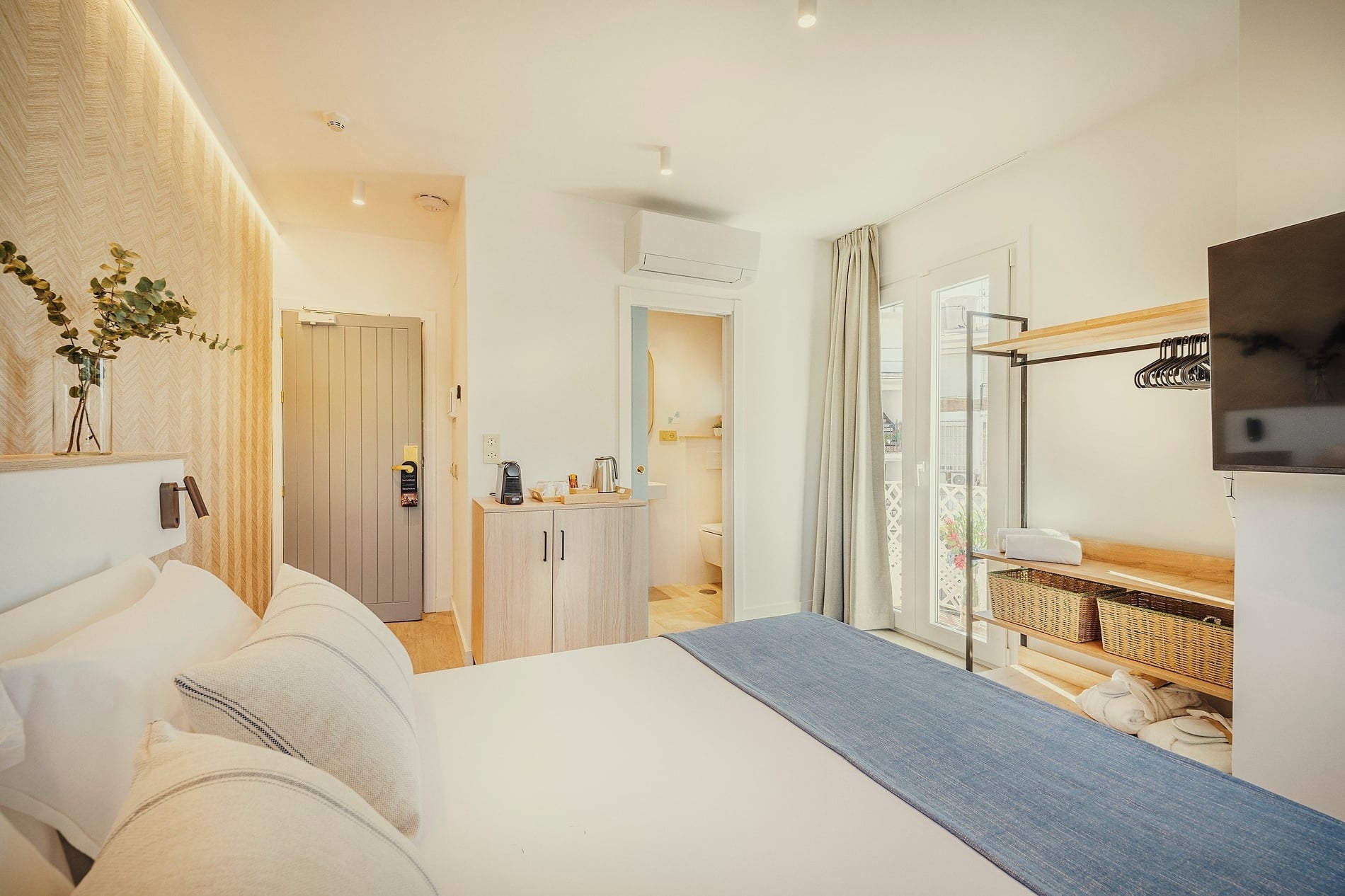 Esta es una luminosa y moderna habitación de hotel con una cama hecha, un armario abierto, un balcón y un baño visible en el fondo.