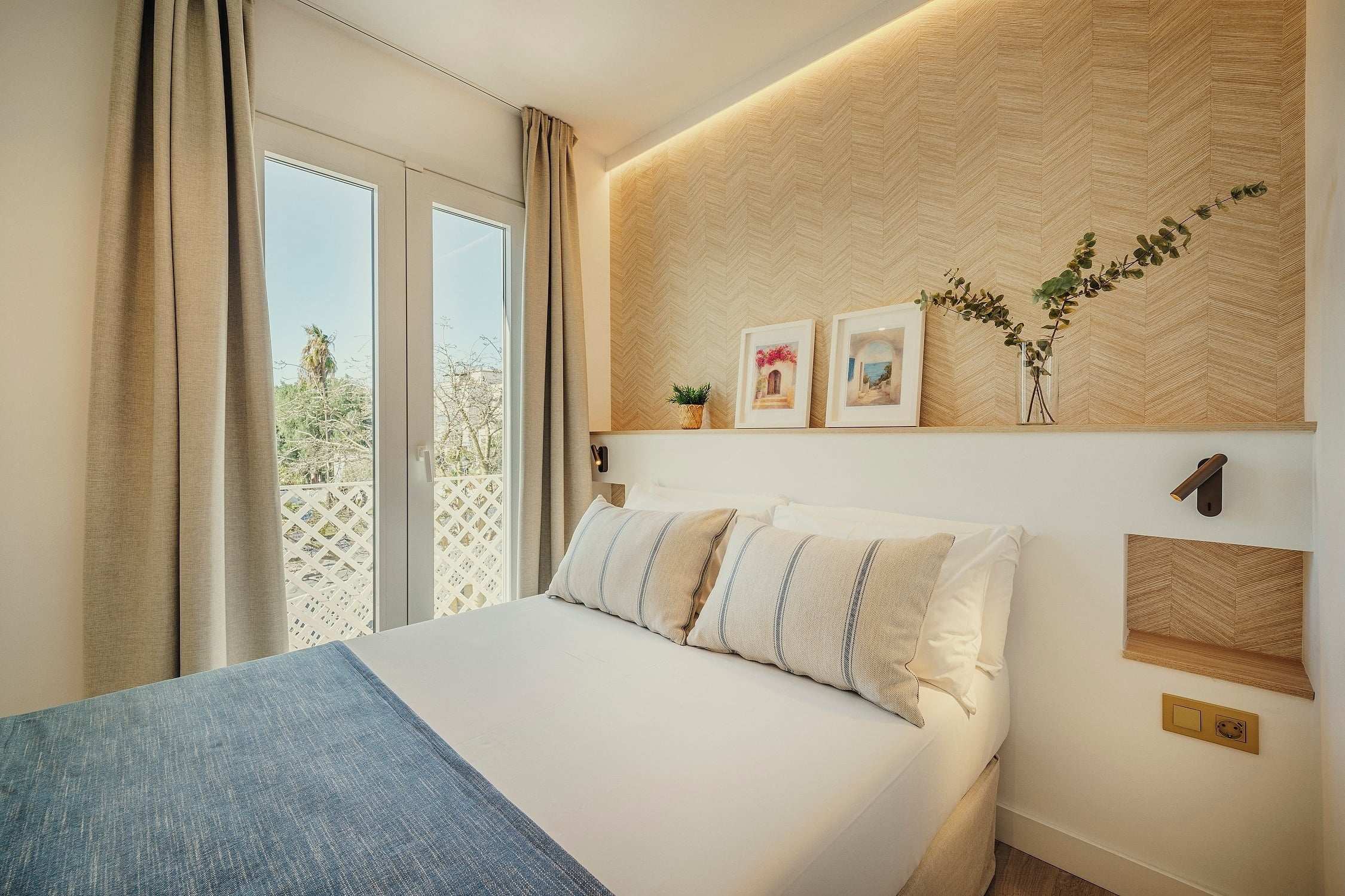 Una habitación moderna y luminosa presenta una cama con ropa de cama blanca y una manta azul, un cabecero de madera con patrón de espiga, una repisa decorada con plantas y cuadros, iluminación integrada y acceso a un balcón a través de puertas acristaladas con cortinas claras.