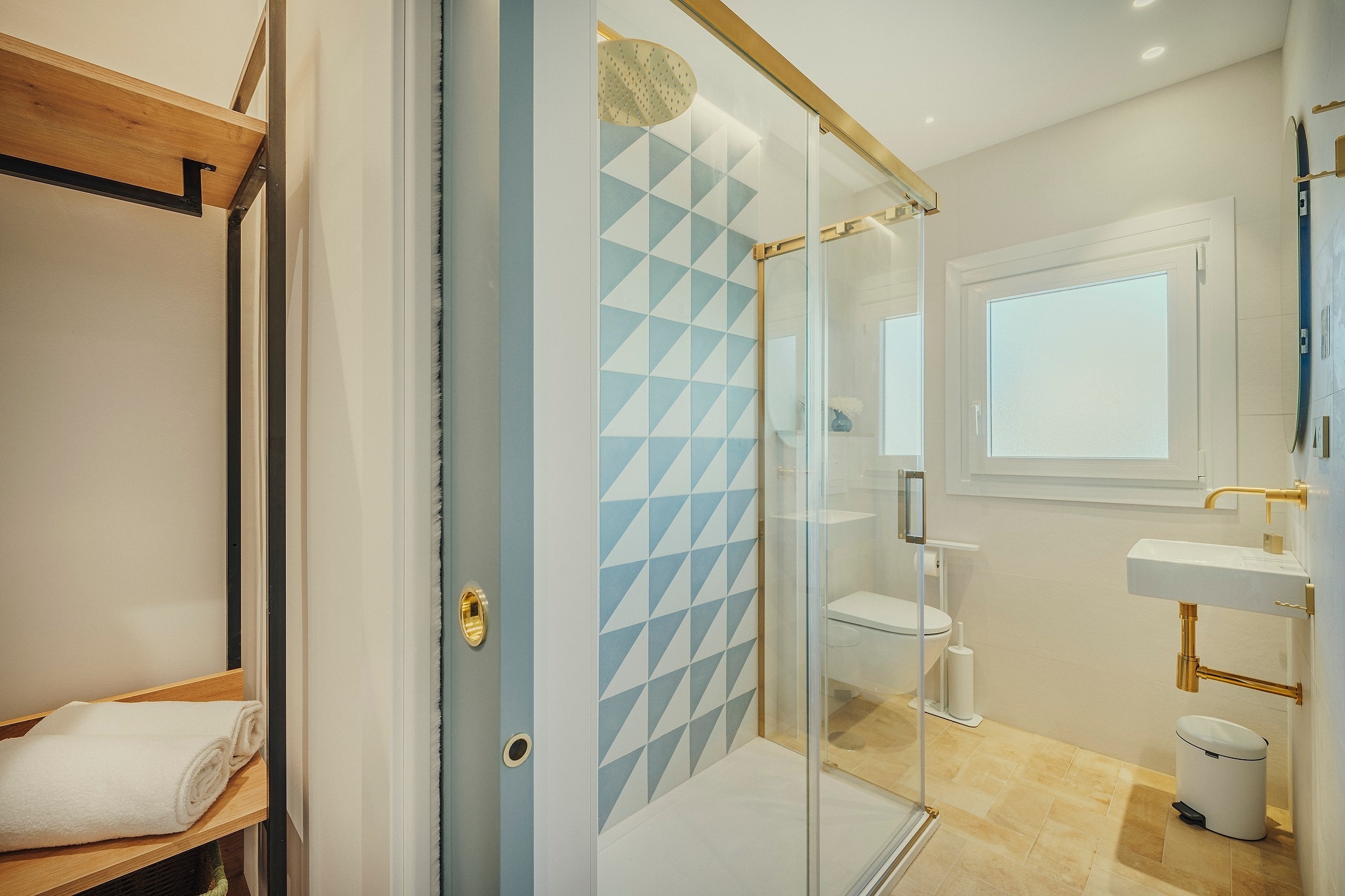 Un baño moderno y luminoso con una ducha de cristal y azulejos geométricos azules, toques dorados, un lavabo y estanterías de madera.