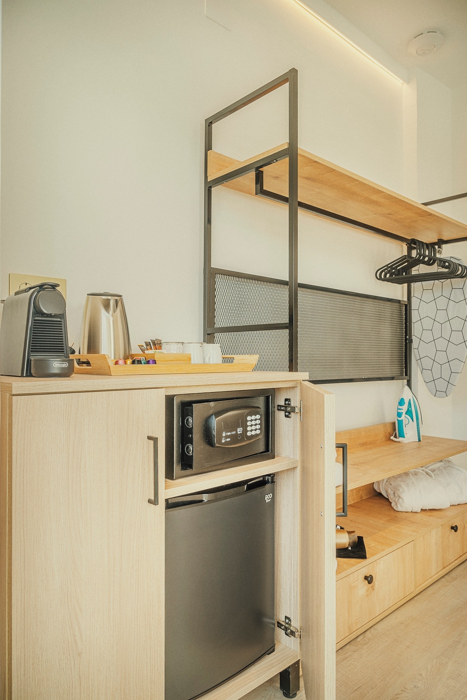 Un rincón de habitación moderno y funcional que incluye una cafetera, un hervidor, una caja fuerte, un minibar y un sistema de estanterías abierto con una tabla de planchar y una plancha.