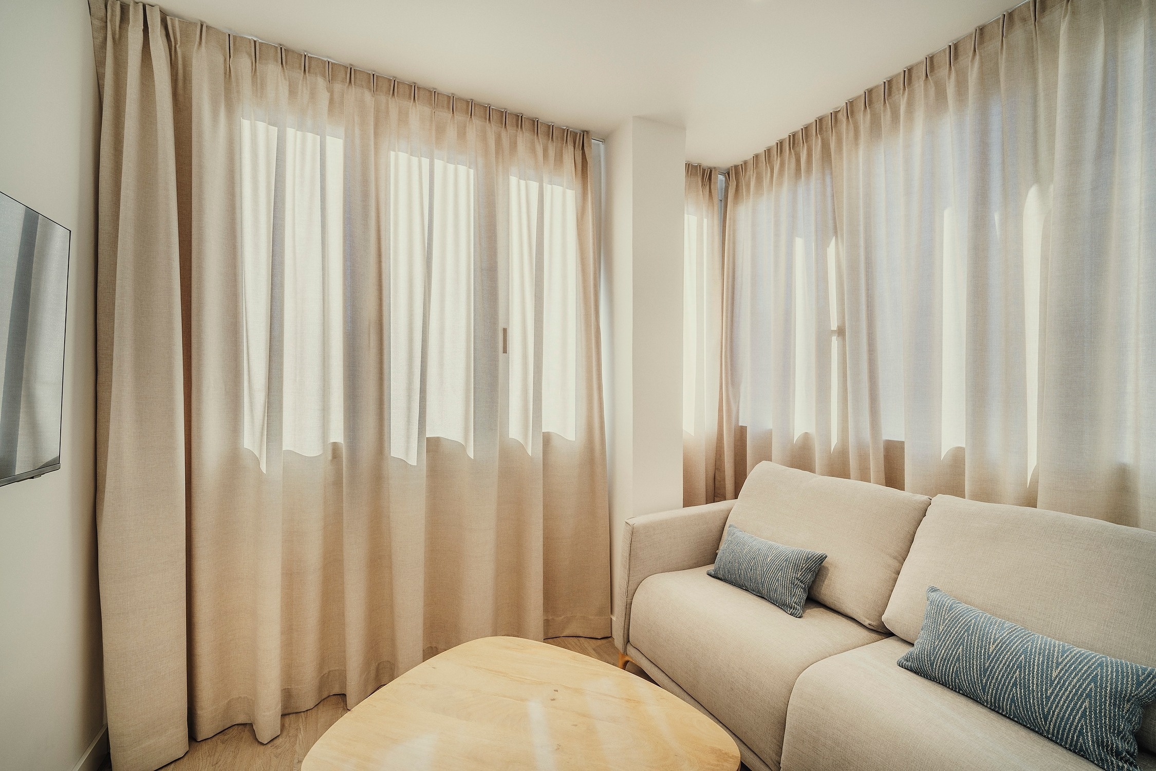Un salón moderno y luminoso con un sofá beige, una mesa de centro de madera y grandes ventanales cubiertos por cortinas traslúcidas.