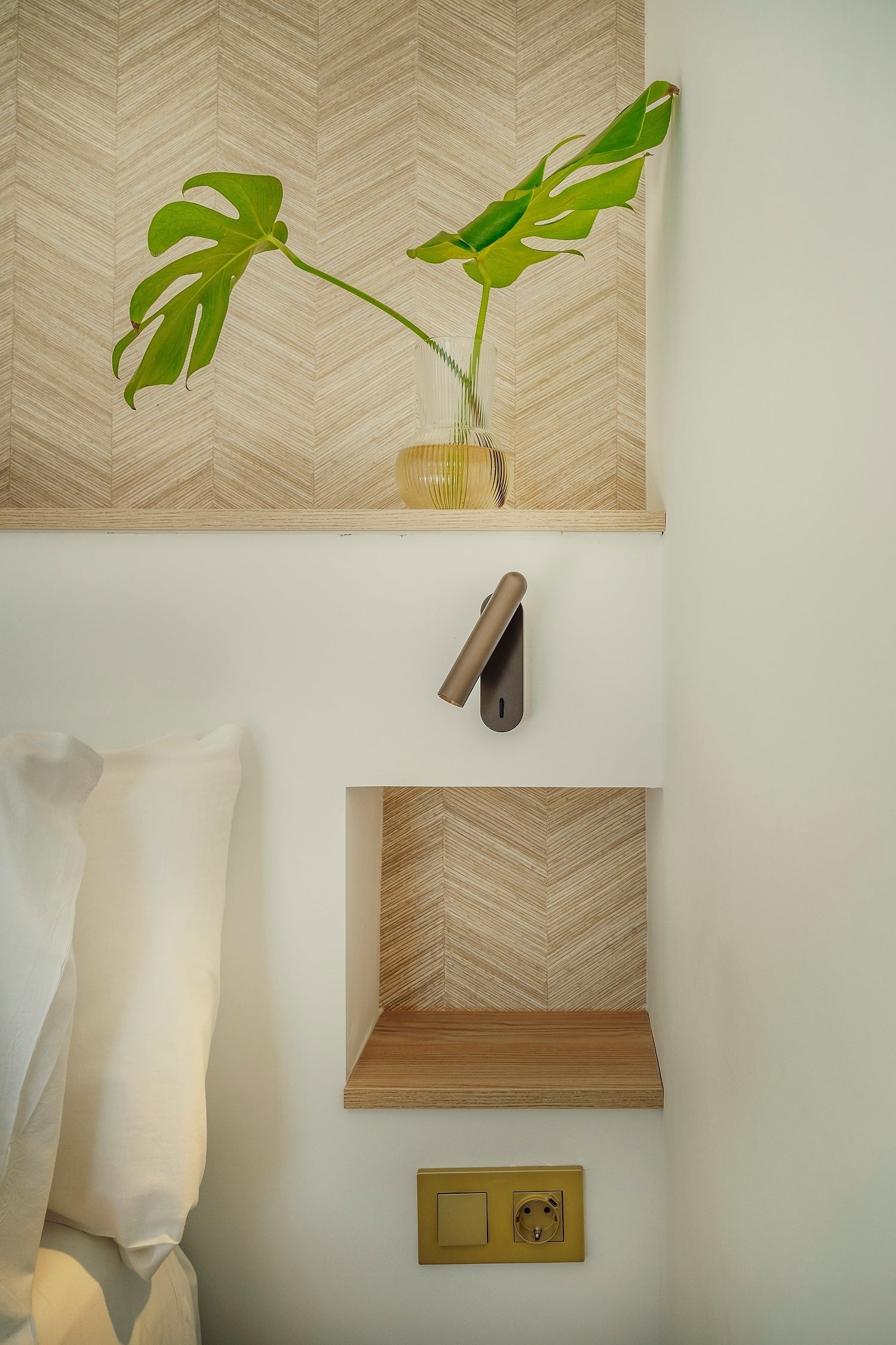 Detalle de una mesita de noche moderna con una hoja de monstera en un jarrón, una lámpara de pared y un enchufe, sobre un fondo de pared con un patrón de espiga de madera.