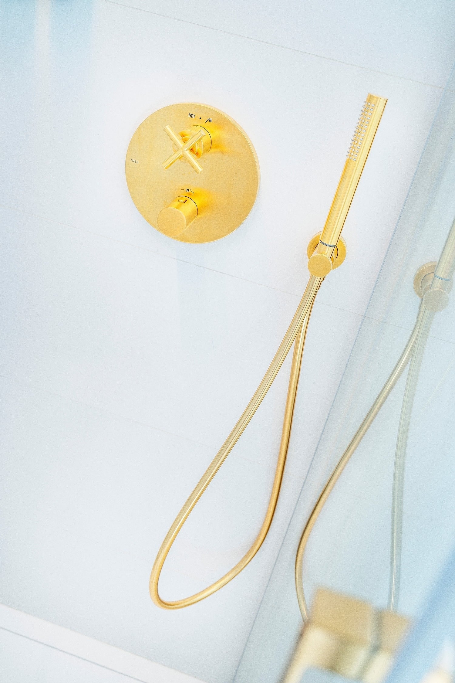 Un grifo y una ducha de mano dorados resaltan en una pared de azulejos blancos, con un reflejo en un panel de vidrio.