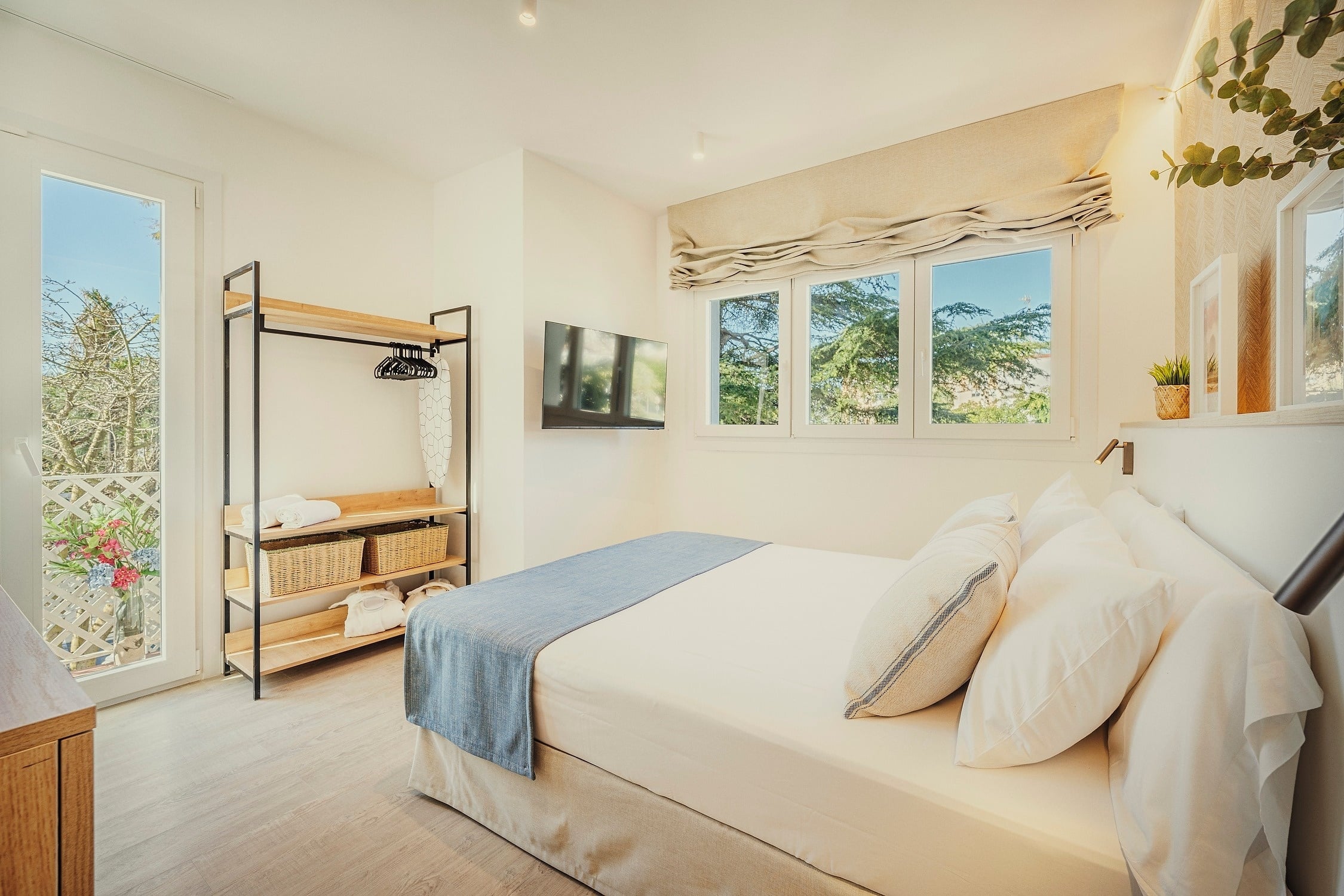 Un dormitorio moderno y luminoso presenta una cama, televisión montada en la pared, estante de ropa minimalista y un balcón con vistas al exterior.