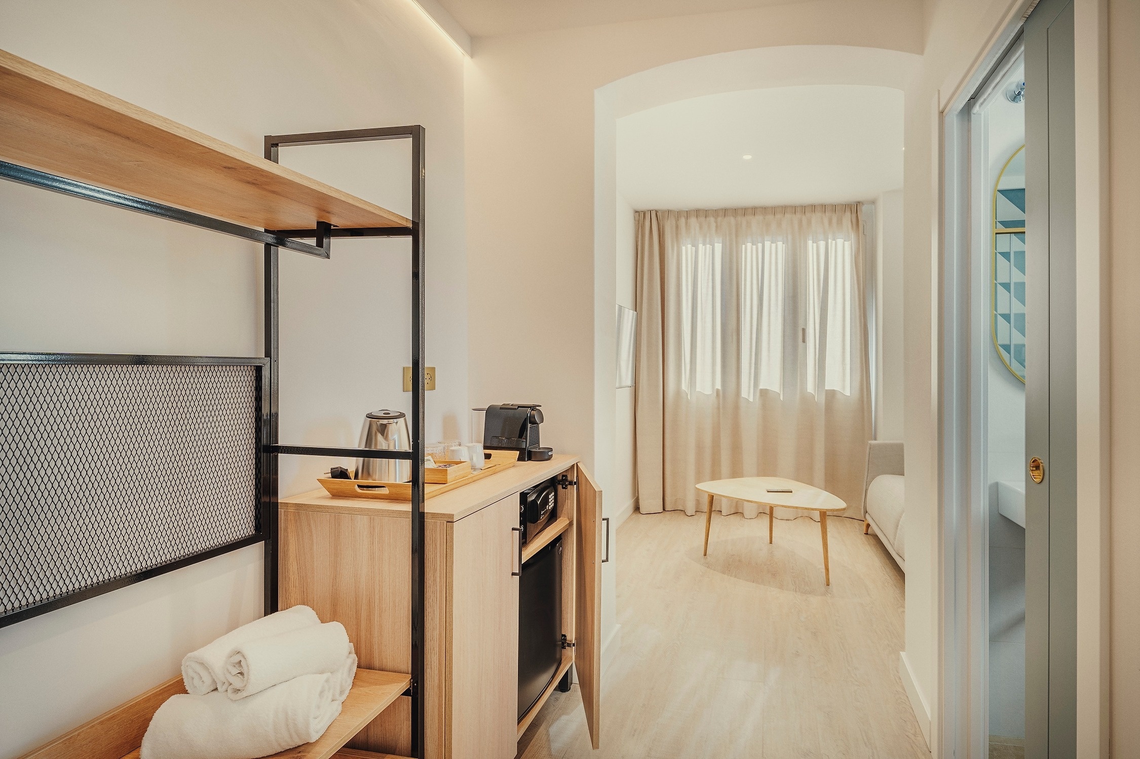 Un interior de habitación moderna muestra una estantería con cafetera y mini-bar, un área de estar con sofá y cortinas, y una puerta entreabierta que revela un baño.