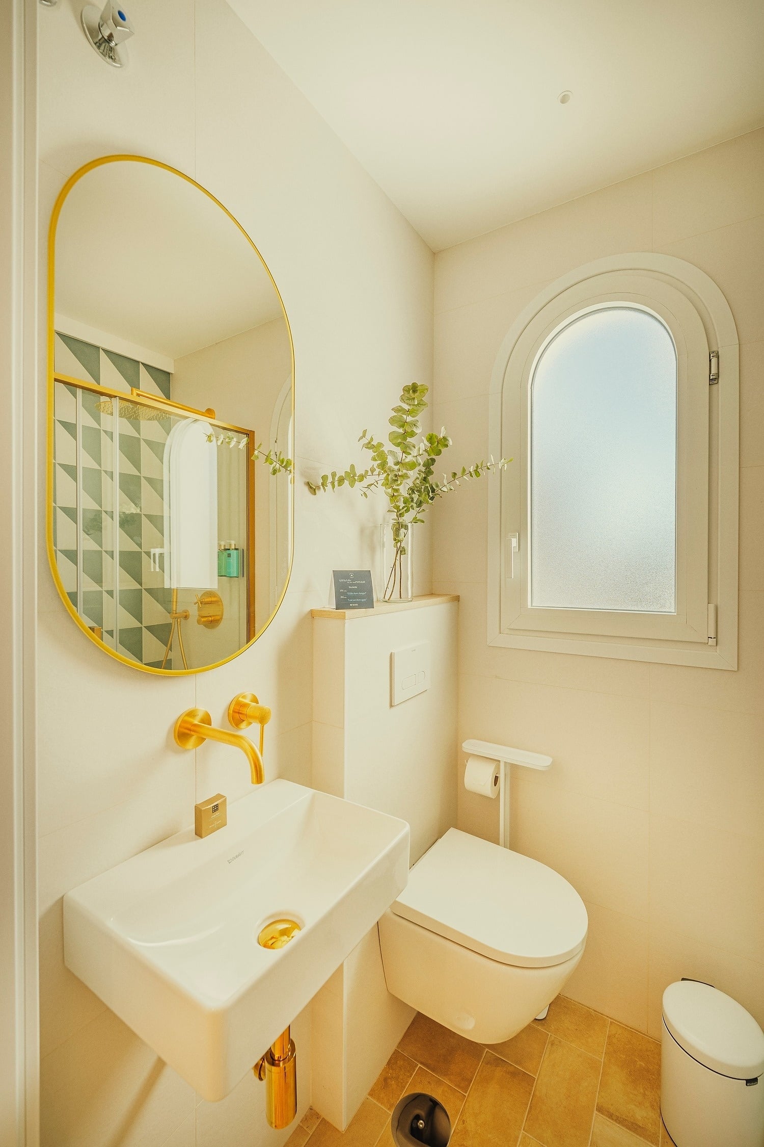 Un moderno y luminoso cuarto de baño con lavabo y grifería dorada, un espejo ovalado, un inodoro suspendido y una ventana arqueada.