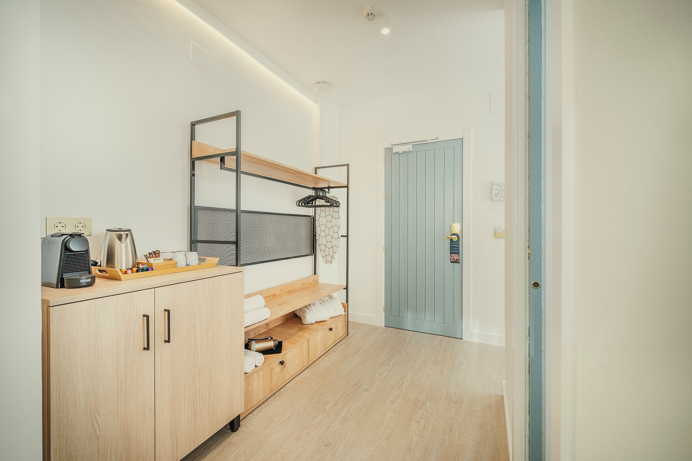 Una moderna y luminosa entrada de una habitación, con muebles de madera clara que incluyen una estación de café y estanterías, junto a una puerta azul claro.