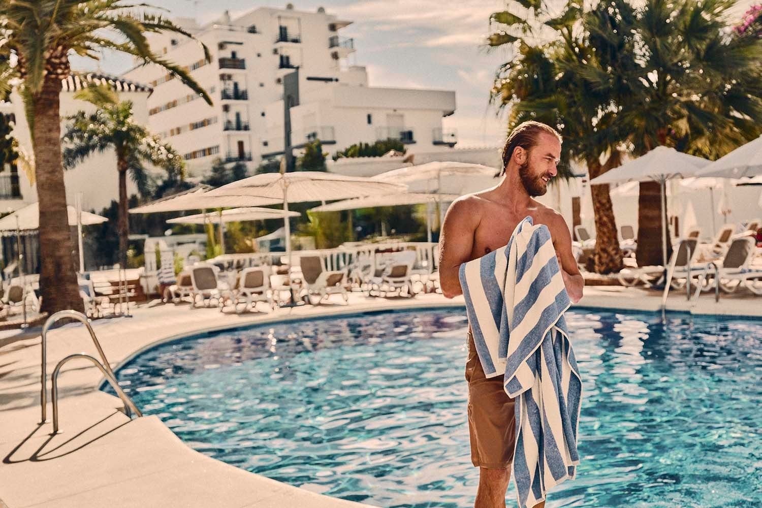Hombre sonriente con toalla a rayas azules junto a la piscina. Villa Flamenca | Beneste Hoteles. Web oficial.