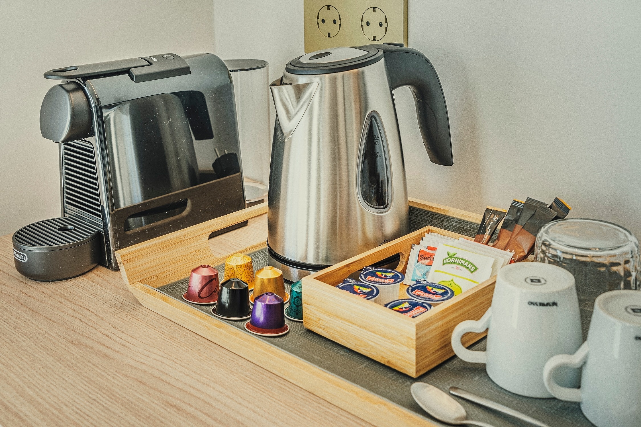 Una estación de café y té que incluye una cafetera Nespresso, un hervidor, cápsulas de café, bolsitas de té y tazas, dispuesta en una bandeja de madera.