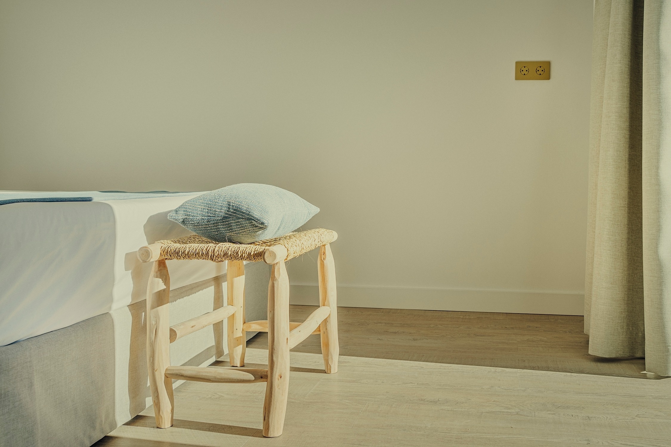 Una habitación soleada con una cama, un taburete rústico de madera con un cojín, un enchufe en la pared y cortinas de tela.