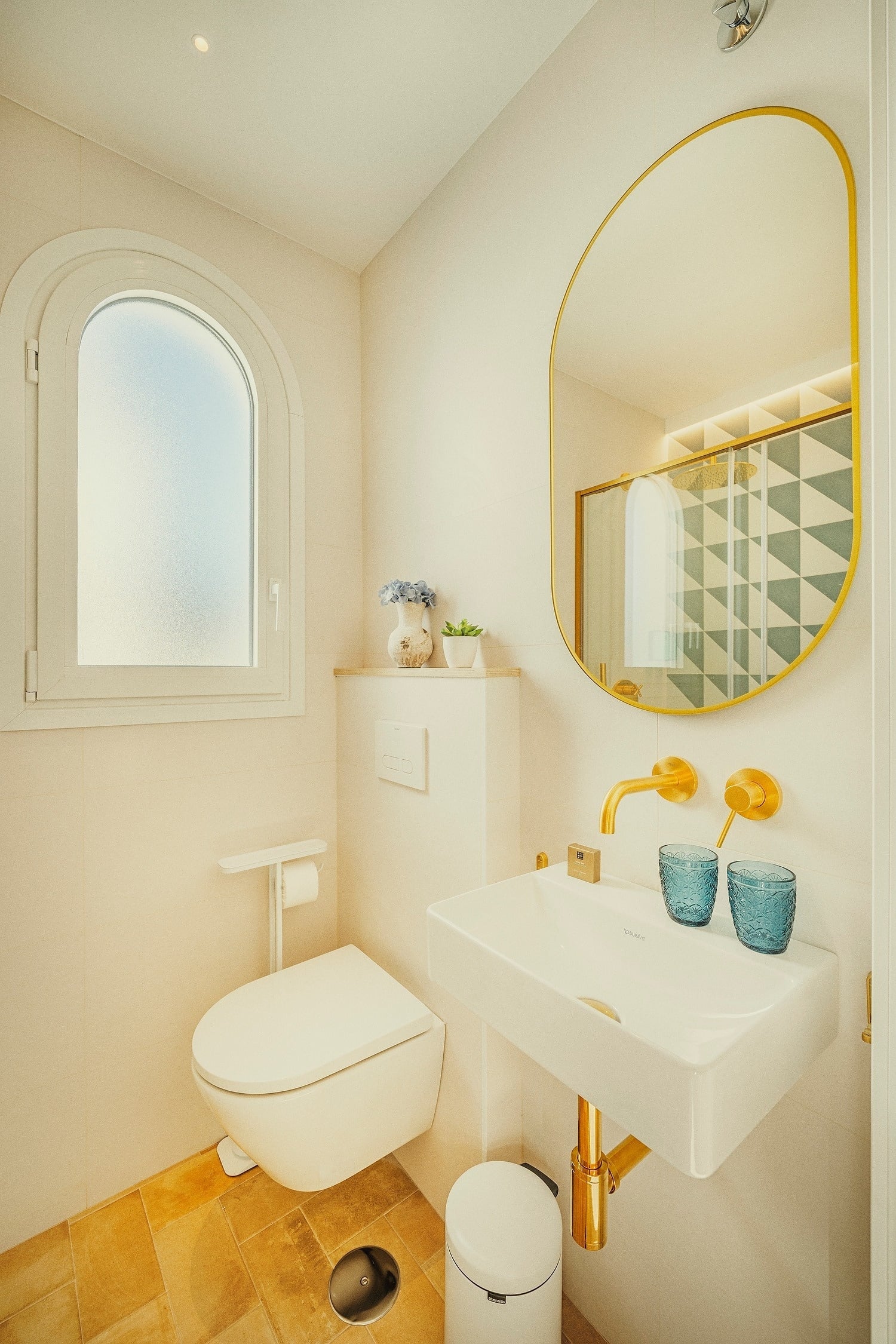 Un baño moderno y luminoso con paredes claras, detalles dorados, una ventana arqueada y un espejo que refleja una ducha con patrón geométrico.