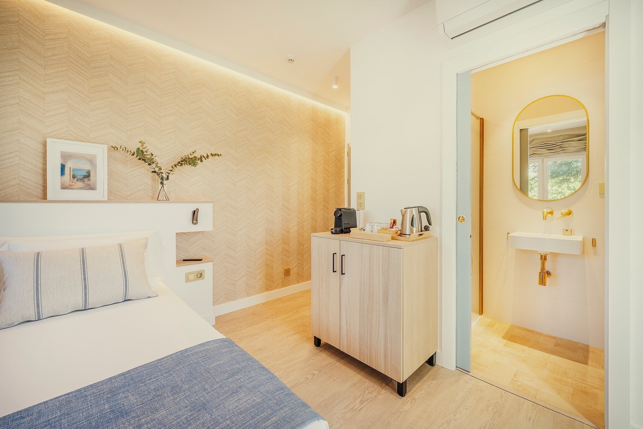 Una moderna y luminosa habitación de hotel con cama, un mueble con cafetera y tetera, y un baño adyacente con lavabo y espejo.