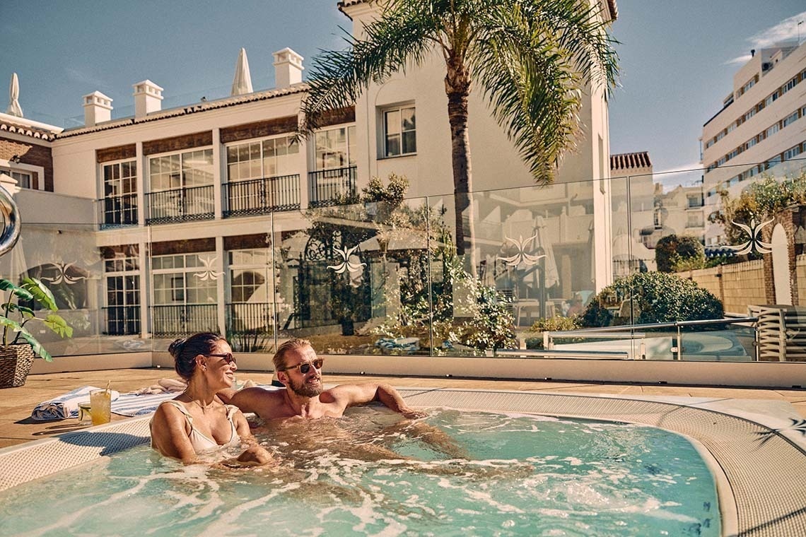 Una pareja sonriente y relajada disfruta de un jacuzzi al aire libre con un edificio blanco y una palmera de fondo bajo un cielo despejado.
