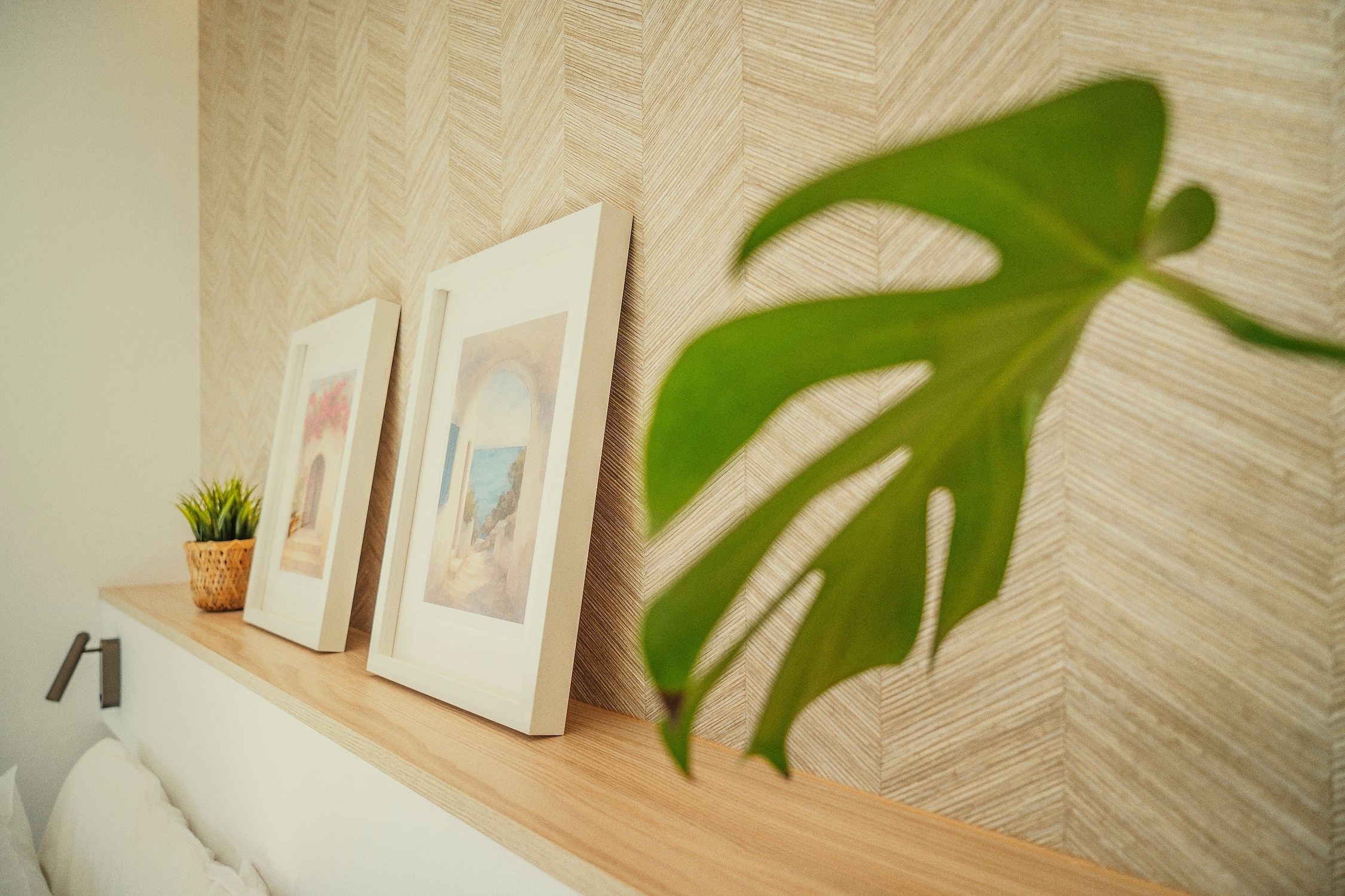 Un estante de madera exhibe cuadros y una pequeña planta frente a una pared con papel tapiz de espiga, con una hoja verde de planta en primer plano desenfocado.