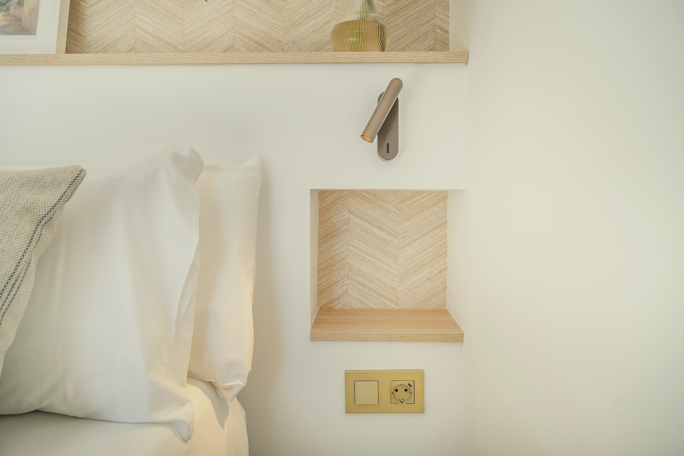 Moderno rincón de dormitorio con estantes de madera clara de espiga, lámpara de lectura y enchufe, junto a almohadas blancas.