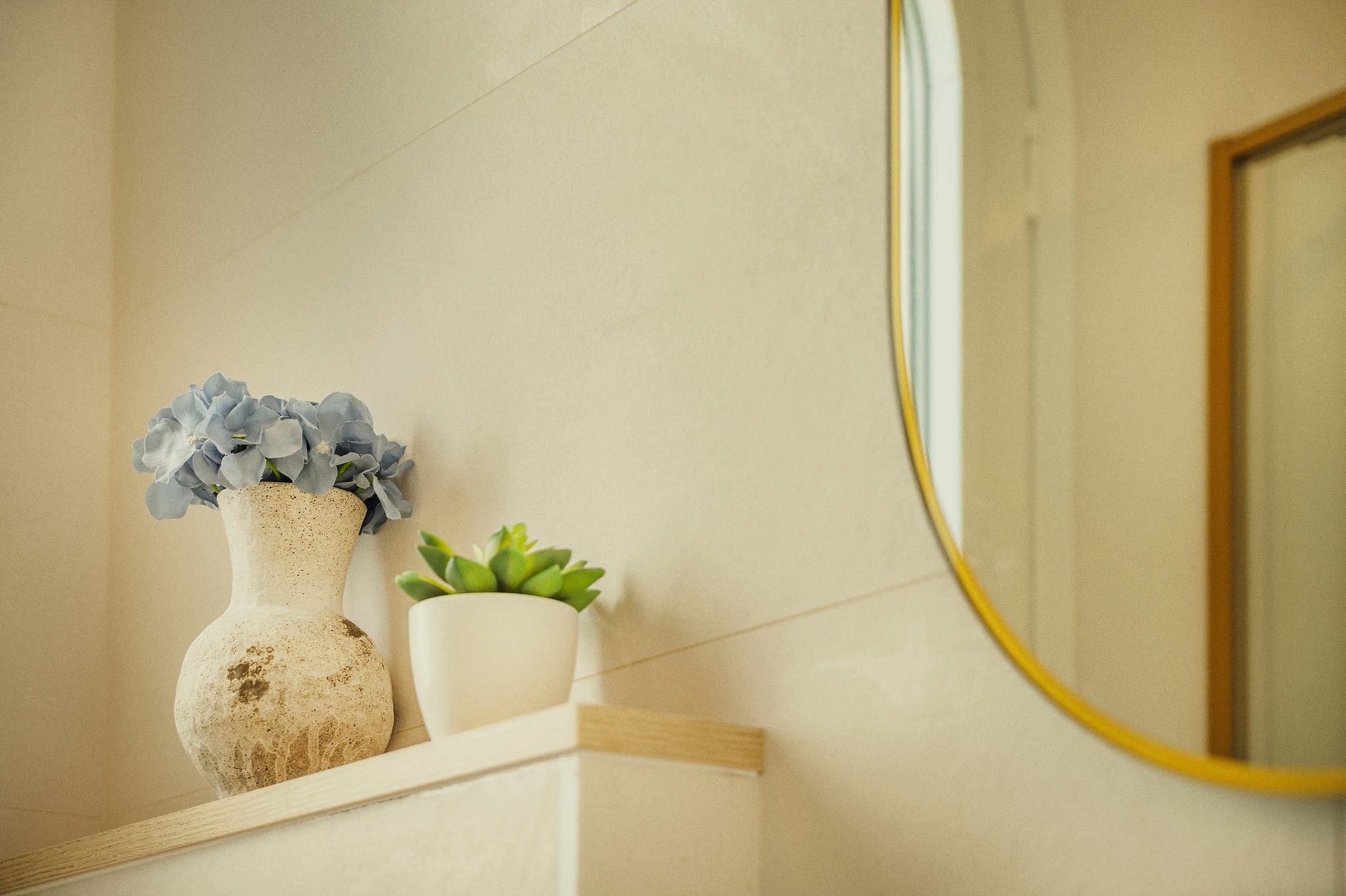 Un jarrón rústico con hortensias azules y una planta suculenta en maceta decoran una estantería clara junto a un espejo redondo de marco dorado parcialmente visible.