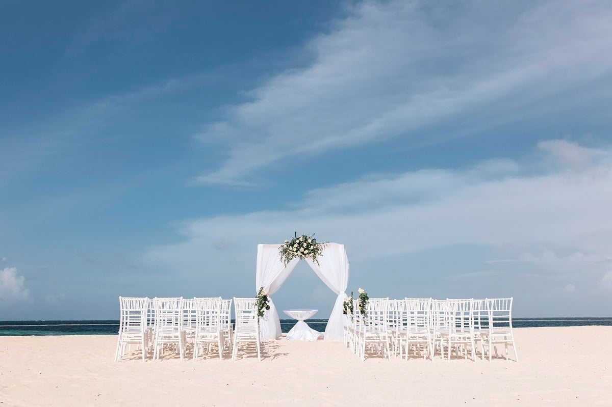 una fila de sillas blancas en una playa con un arco decorado con flores