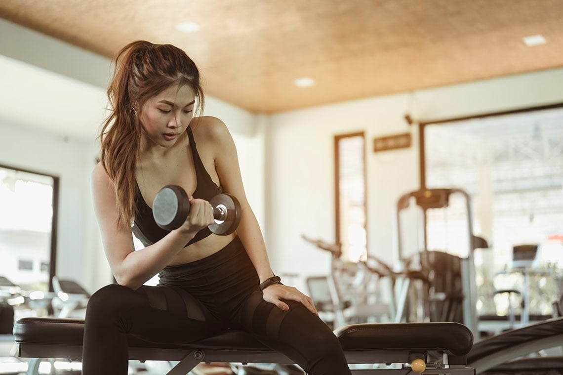 una mujer sostiene una mancuerna mientras está sentada en un banco en un gimnasio