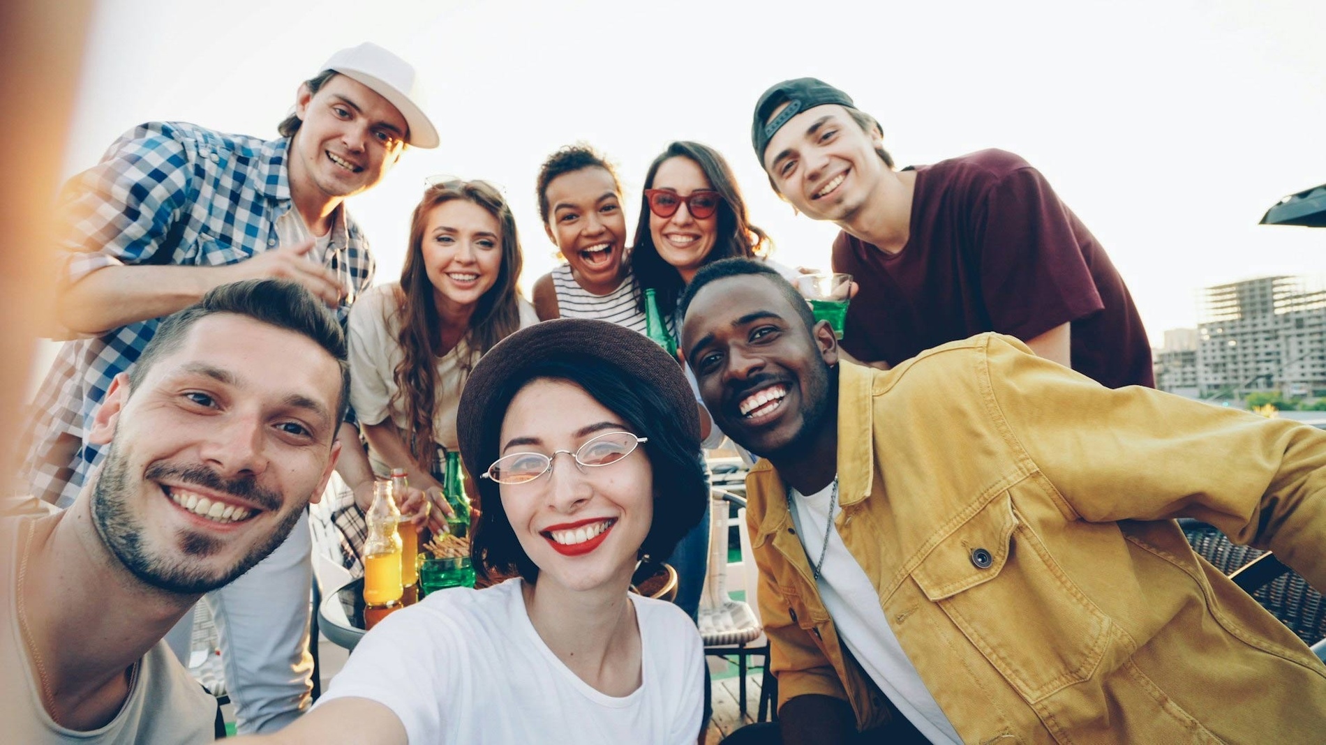 A diverse group of eight young adults joyfully take a group selfie at an outdoor gathering with drinks.