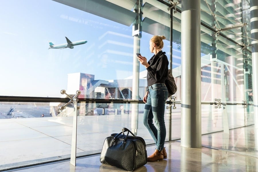 Une femme tire sa valise dans un terminal d'aéroport moderne, avec un avion visible sur le tarmac à travers les grandes baies vitrées.