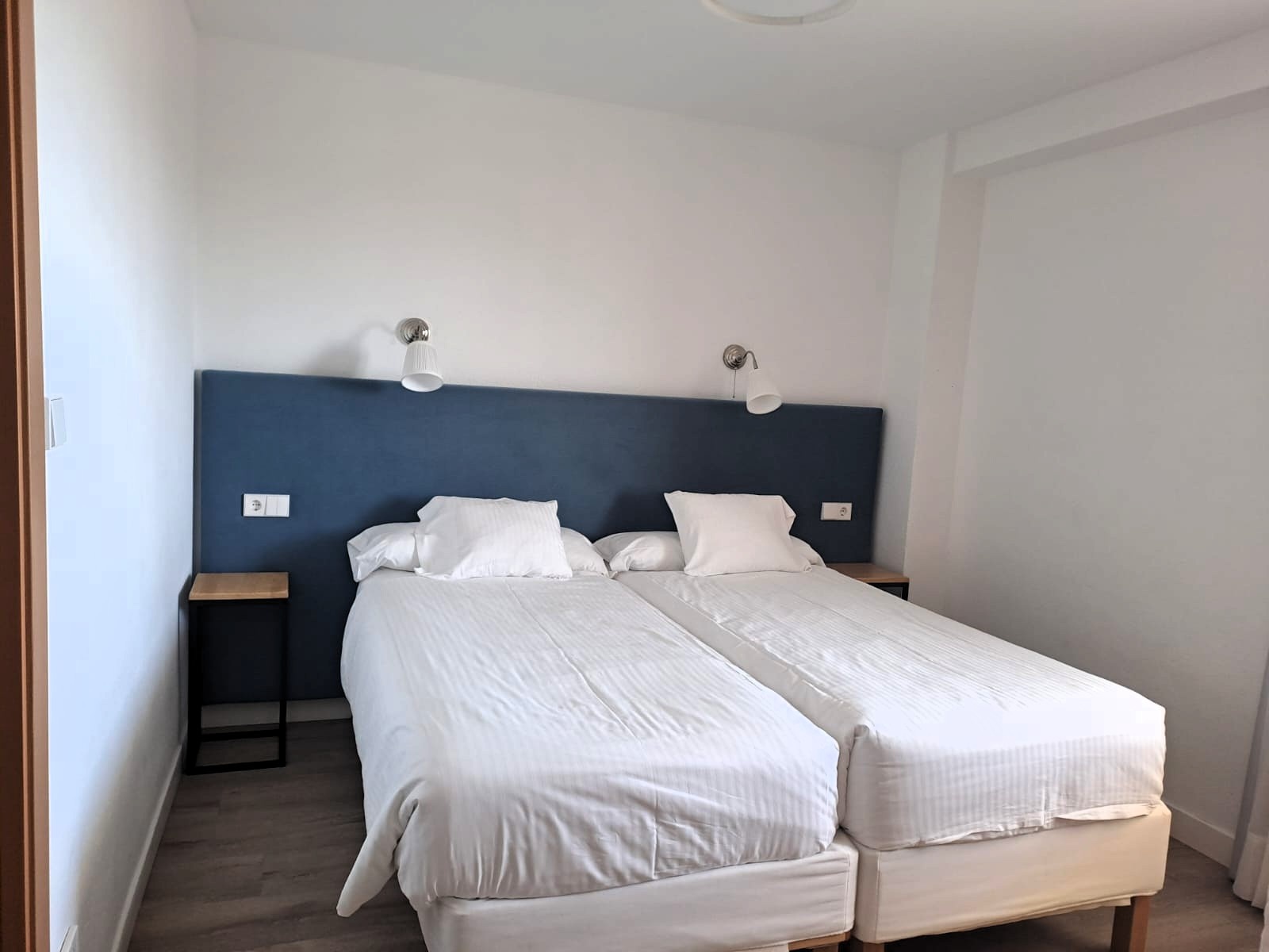 Una habitación de hotel sencilla con dos camas individuales vestidas de blanco, un cabecero azul, paredes blancas y suelo de madera.